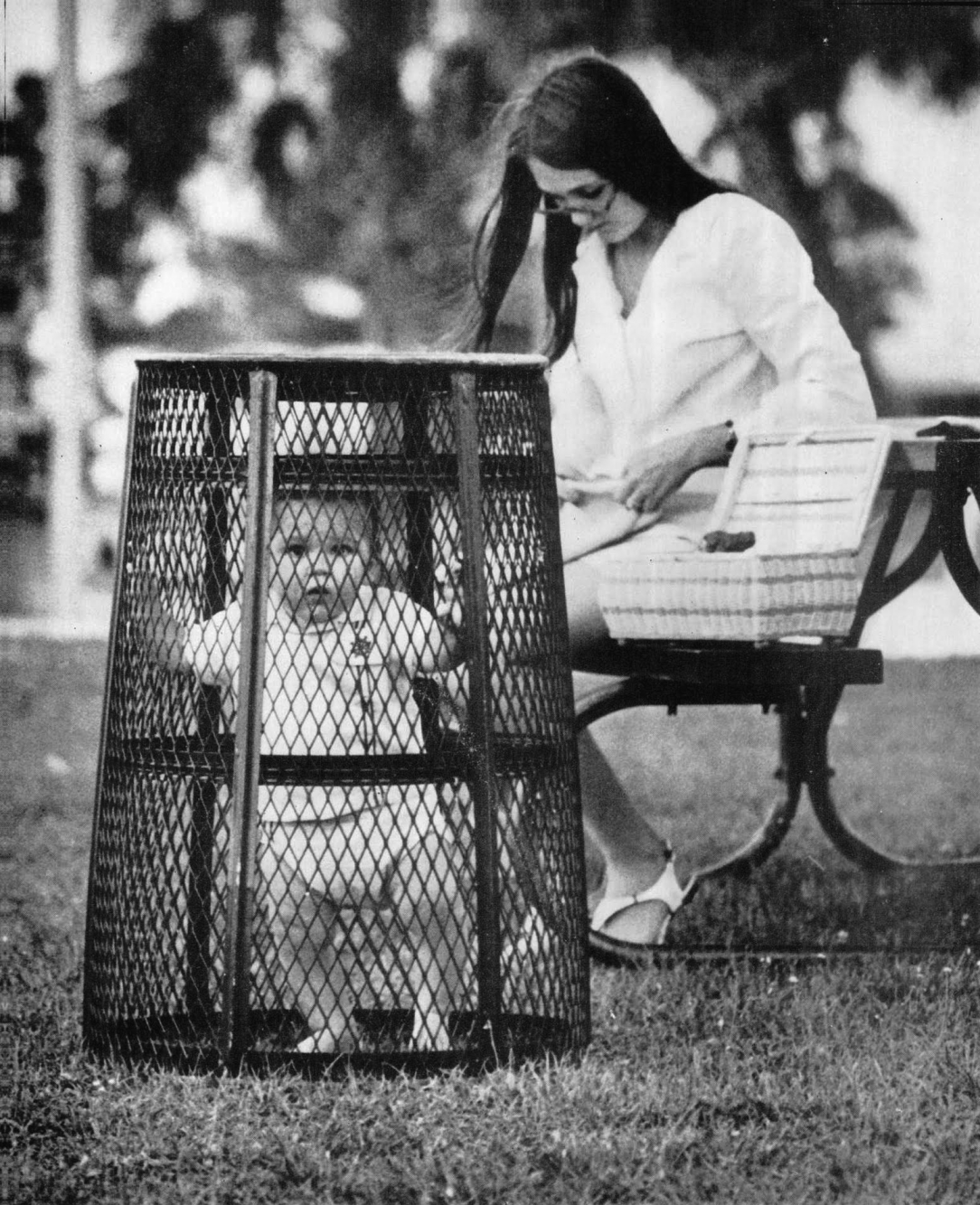 A mom uses a trash can to contain her baby while she crochets in the park, 1969